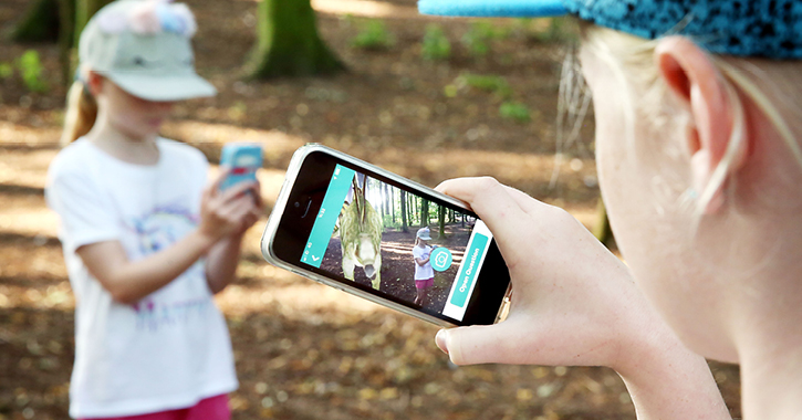 two girls playing with the Love Exploring app in a country park showing the dinosaur hunting app on mobile phone screen.